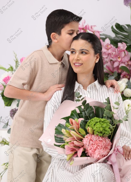 Image of a woman wearing a striped shirt holding a bouquet of mixed flowers, including pink lilies and green blooms. A young boy in a beige shirt stands beside her, kissing her forehead. The background is adorned with pink flowers and green foliage.