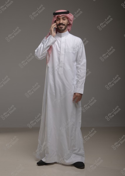 A man wearing a traditional white thobe with a red checkered shemagh and a black agal, standing and talking on the phone with a smile. He appears to be in a studio against a gray background. The attire suggests he might be from Saudi Arabia.
