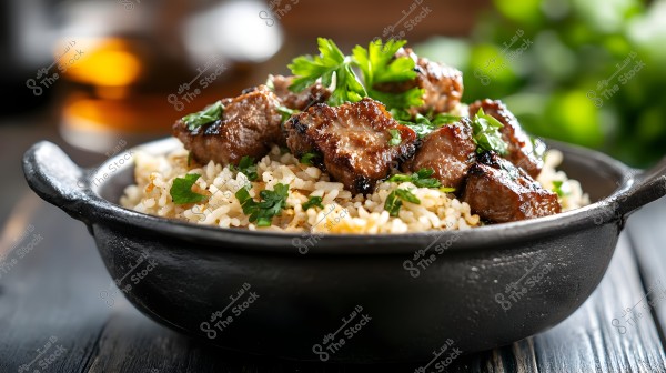 Image of a dish containing grilled meat pieces on a bed of cooked rice, garnished with fresh parsley. The dish is placed in a black bowl and set on a dark wooden surface. In the background, there are blurred colors of green and orange.