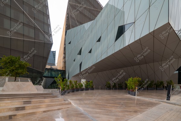 Image of a modern building with a contemporary architectural design featuring glass and metal facades. Geometric lines are visible on the facades and different angles. On the left side, there is a small tree on a short platform, and on the right, there are plants in pots. A smooth stone entrance leads inside.