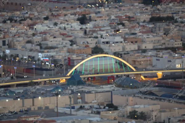 منظر جوي لمدينة يظهر فيها جسر ذو قوس مضيء بألوان زرقاء وصفراء, ويقع في وسط منطقة سكنية مليئة بالمنازل البيضاء والمباني ذات الطراز الحديث. الطريق السريع يمر تحت الجسر, ويوجد عدة لوحات مضيئة للمحلات التجارية على جانبيه.