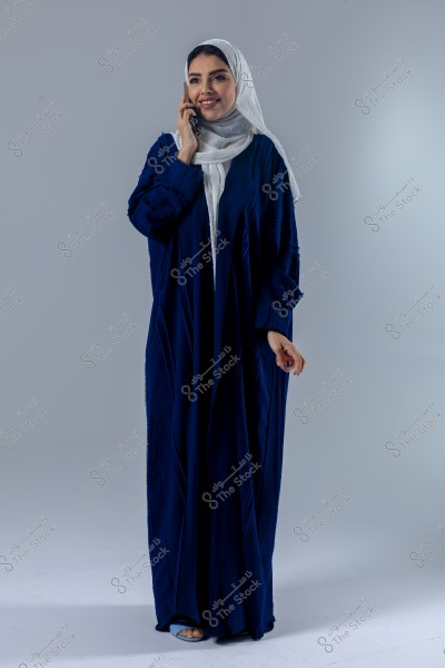 Image of a woman wearing a blue abaya and a white hijab, smiling while talking on the phone. She appears to be of Saudi or Gulf origin. The background is a simple gray.