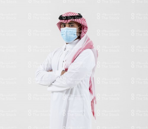 شاب سعودي يرتدي الزي السعودي الرسمي على خلفية بيضاء وهو واضع الكمامة على وجهة.