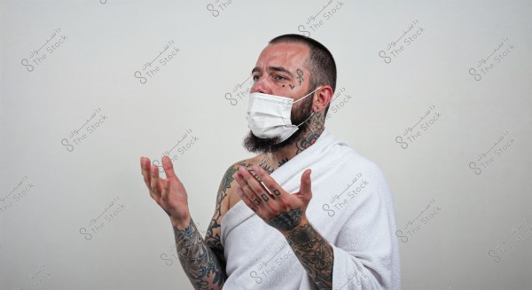 An image of a man standing against a white background, wearing a medical face mask and wrapped in a white cloth that resembles Ihram. His arms and part of his face are covered in tattoos. His hands are slightly raised, as if in a praying position.