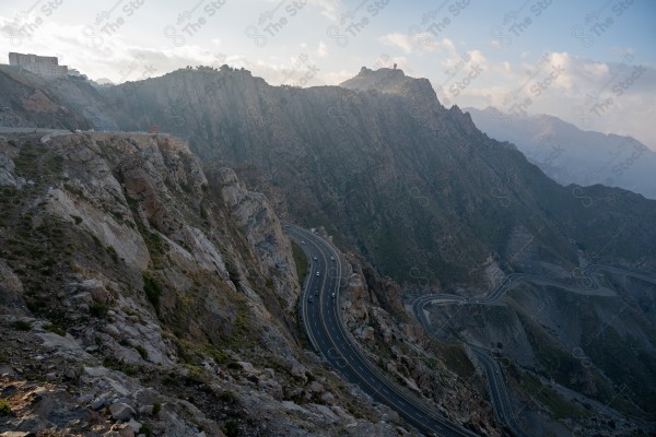 A snapshot of a series of towering rocky mountains and paved roads extending in the Al Hada Mountains in Taif, showing a cloudy sky during the day, mountain heights, Taif Mountains, mountain range