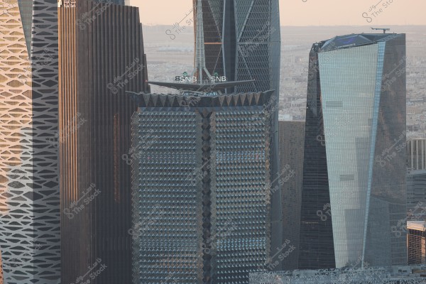 An image of skyscrapers in Riyadh, Saudi Arabia, showcasing modern and luxurious architectural designs. In the foreground, there is a tower with a distinctive geometric design covered in glass that beautifully reflects light, surrounded by other towers with glass facades and varied shapes.