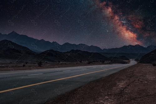 A paved road in the middle of rocky mountains and the sky appears clear, sparkling with stars and planets at night.