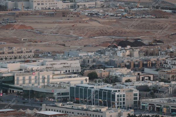 The image shows an aerial view of an urban area in Saudi Arabia, featuring a cluster of modern residential buildings and commercial offices. The photo also includes a large construction site with heavy machinery excavating and moving soil. In the foreground, there is a commercial complex, residential units, and other buildings surrounding the site.