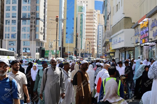 A crowded street with many people wearing traditional pilgrimage attire such as white robes and abayas. Modern buildings are visible in the background in an urban area. Shop signs are visible on the right side of the image.