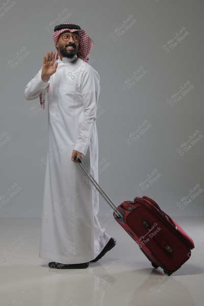 A man wearing traditional Saudi attire, including a white thobe, a red checkered ghutra, and a black agal. The man is smiling and waving, while pulling a red suitcase with a telescopic handle. The background is gray.