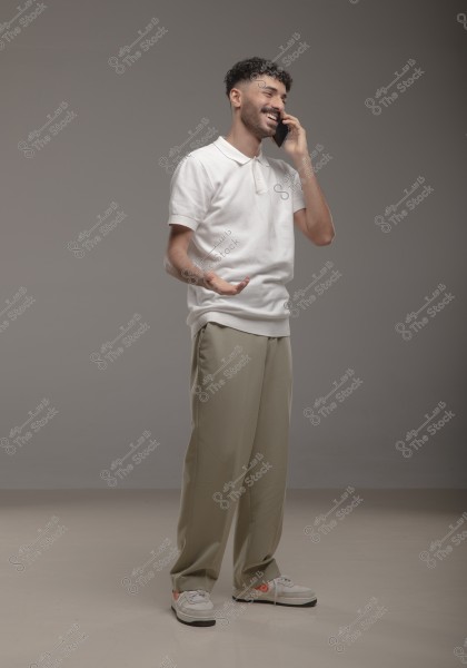 An image of a man wearing a white polo shirt, beige pants, and sneakers, talking on the phone with a smile on his face. He is standing in an indoor setting with a gray background.