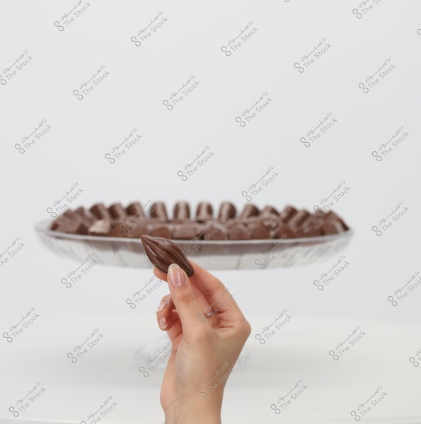 A hand holds a cocoa bean-shaped piece of chocolate in front of a large glass dish filled with similar chocolate pieces. The background is white, and the hand appears at the bottom of the image.