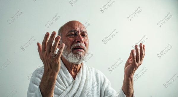 Portrait of an elderly man raising his hands towards the sky, appearing sad with tears on his face. He is wearing a white robe that resembles an Ihram garment. The background is white and serene.