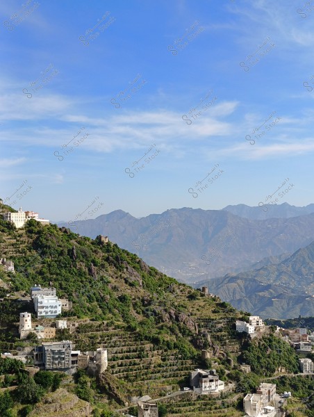 A scenic view depicting a mountainous village situated on green, terraced hillsides. The houses are constructed in traditional styles, with some featuring round towers. In the background, distant mountains are clearly visible under a mostly clear blue sky with some light clouds.