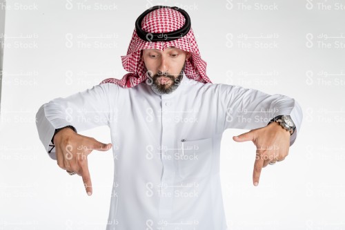 بورتريه رجل سعودي يقف على خلفية بيضاء، يرتدي الثوب، يقف امام الكاميرا ويقوم بعمل حركات بيديه، يمد يديه نحو الكاميرا ويشير بأصابعه نحو الاسفل