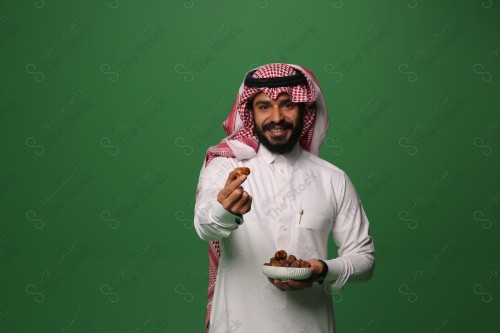 بورتريه رجل سعودي يرتدي الزي السعودي التقليدي يحمل طبق من التمر ويقدم تمرة لأحدهم