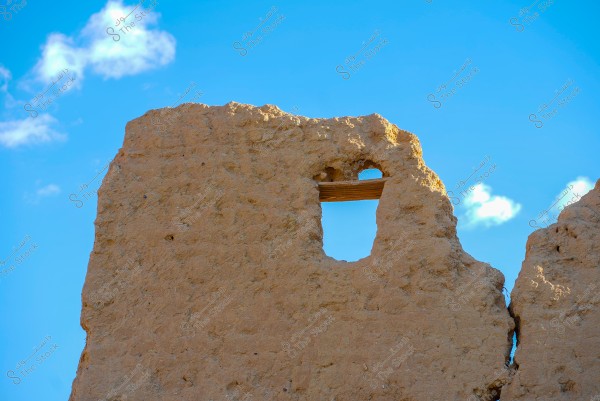 تظهر الصورة جدارًا طينيًا قديمًا مع فتحة نافذة صغيرة, يتخللها قطع خشبي, يتماشى مع لون الجدار. خلفية السماء زرقاء مع وجود بعض السحب البيضاء الخفيفة.