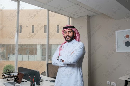 رجل سعودي يرتدي الزي السعودي التقليدي يقف مكتوف اليدين أثناء إجتماع في العمل نهارًا