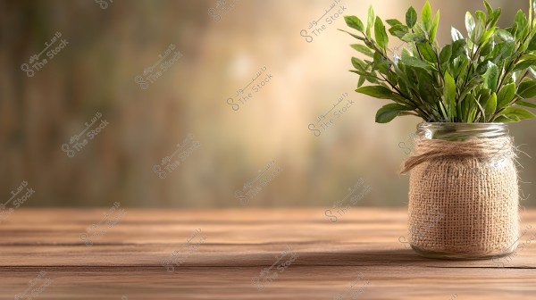 Image of a glass jar wrapped with a piece of burlap, containing a bunch of green branches with lush leaves. The jar is placed on a wooden surface with a blurred background in shades of brown and gold.