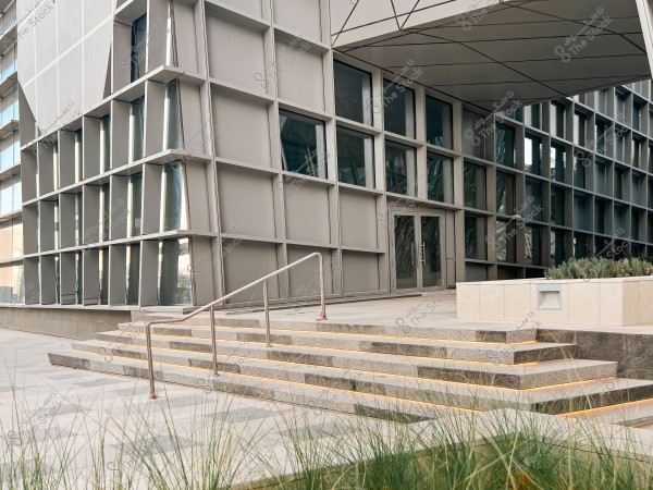 The image shows a modern-designed building with a distinct architectural facade consisting of glass windows and a metal framework. In the foreground, there are marble steps with a metal railing, accompanied by small plants. The design is characterized by straight geometric lines and sharp angles, reflecting a contemporary style.