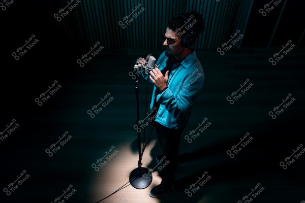 شخص يرتدي نظارات شمسية وسماعات رأس واقف أمام ميكروفون في استوديو تسجيل مظلم.