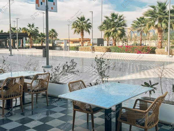An image showing an outdoor area of a café, with wicker tables and chairs set on a black and white checkered floor. In the background, there is a wide walkway surrounded by palm trees and flowering plants. The weather is sunny and the sky is blue with some clouds.