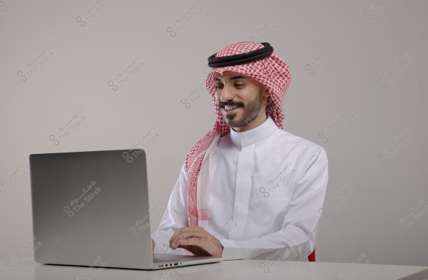 صورة لرجل يرتدي ثوبًا أبيض وغترة حمراء مع عقال أسود يجلس أمام حاسوب محمول. يبدو الرجل مبتسمًا ويستخدم الكمبيوتر. الخلفية ذات لون رمادي فاتح.