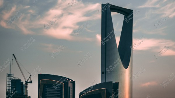صورة لبرج شهير في مدينة الرياض, السعودية, يعرف ببرج المملكة. يظهر البرج بتصميم مميز في شكل القوس, وتحيط به سحب في السماء ذات لون وردي خفيف عند الغروب. يظهر في المقدمة رافعة بناء ومباني أخرى حديثة.
