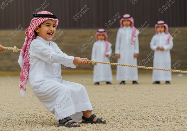 The image depicts a group of children playing tug of war in an outdoor area. In the foreground, a child is sitting on the ground, pulling the rope with enthusiasm, smiling joyfully. The children are wearing traditional Saudi attire, consisting of a white thobe and a red checkered headscarf. In the background, three more children, dressed similarly, are standing and watching the event. The ground is covered with small pebbles.