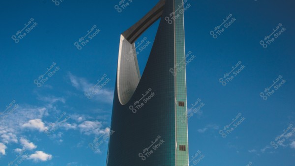 Image of the Kingdom Tower in Riyadh, Saudi Arabia. The tower features a unique design with an inverted arch shape and is covered with glass reflecting the blue sky with a few light clouds.