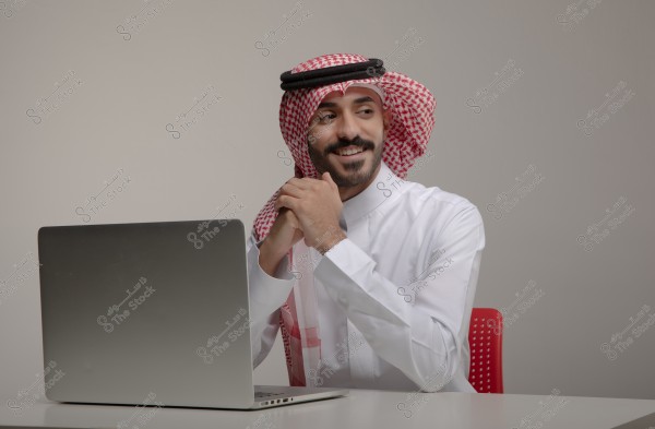 A portrait of a man sitting at a table in front of a laptop. He appears to be wearing a white thobe and a red checkered ghutra with an agal, traditional attire that might indicate Saudi origin. He is smiling broadly and looking to the side, with his hands clasped in front of him. The background is a neutral gray color.