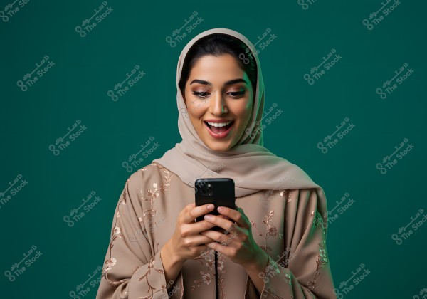 A portrait of a woman wearing a beige hijab and an abaya with floral decorations. She is smiling and looking at a smartphone in her hands. The background is dark green, and the woman appears happy and pleasantly surprised.