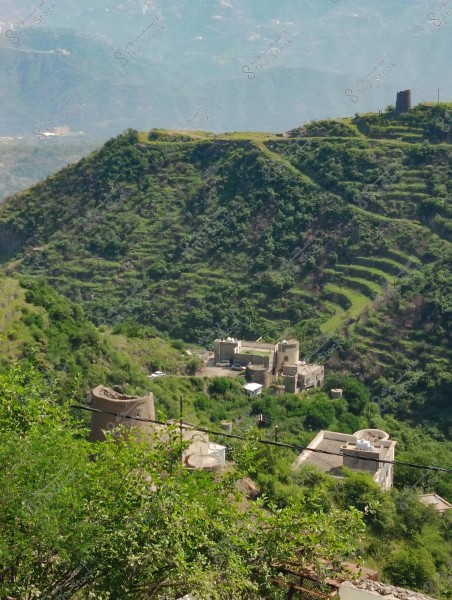 A natural landscape featuring green hills covered with dense vegetation and agricultural terraces. In the foreground, there is a cluster of traditional buildings with flat roofs and small towers, constructed from stone and mud, nestled amidst the greenery. The distant horizon reveals other misty hills.