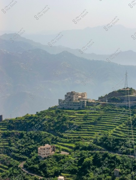 Image showing a natural landscape of a mountainous area covered in green agricultural terraces. The photo features residential buildings perched on the hilltop along with a power line. The background has layers of misty mountains under a clear sky.
