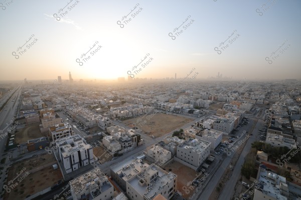 A panoramic view of a large city at sunset. Residential and commercial buildings are scattered in an organized manner, connected by streets. In the background, sunlight gleams behind modern skyscrapers, giving the scene a golden glow. The sky is clear with a few wispy clouds.