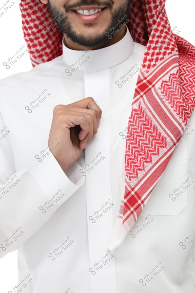 Image of a man wearing a traditional white thobe and a red and white ghutra covering his head. Part of his face is visible with a smile and light beard, and he is holding the edge of the thobe with his hand.