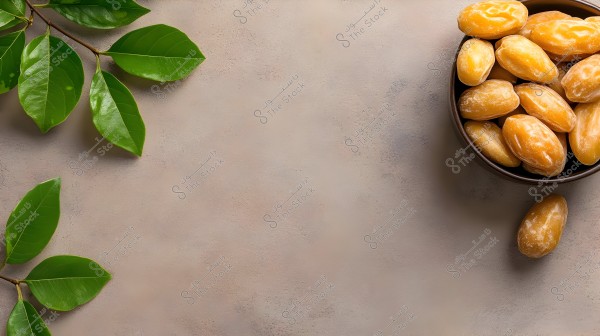 The image shows a group of golden-colored dates placed in a round bowl on a beige surface. On the left side, glossy green plant branches extend across the surface, creating a beautiful contrast between the dates and the leaves.