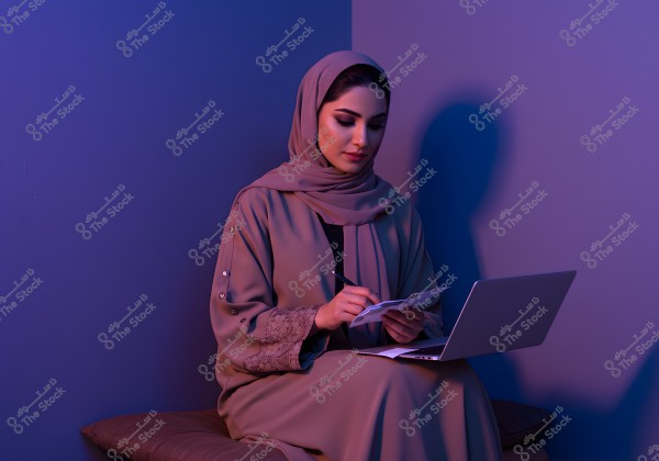 A portrait of a woman sitting in a room with soft blue and pink lighting, wearing a hijab and abaya. She appears to be writing in a small notebook, with an open laptop in front of her. The serene and calming atmosphere suggests a mood of focus and quiet contemplation.