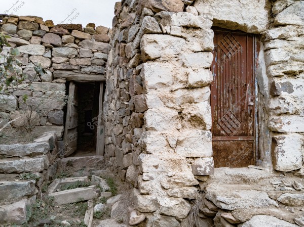 An image showcasing an old stone building with traditional architectural details. The structure features a wall made of naturally stacked stones, with a traditional wooden door leading inside. Wild plants are visible on the surrounding ground, adding a natural beauty to the scene.