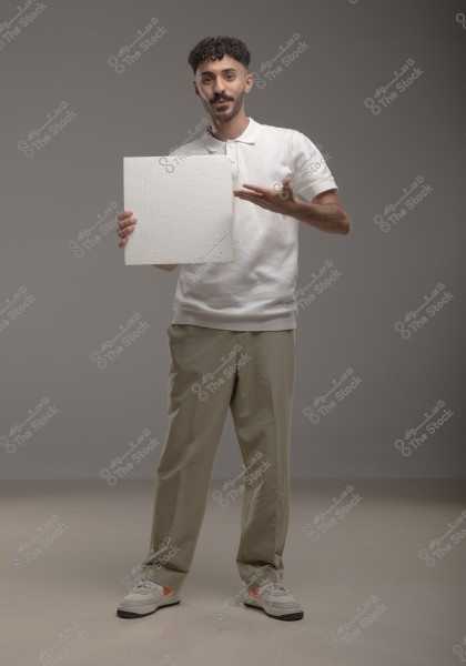 Image of a man standing in a studio with a gray background. The man is wearing a white polo shirt, khaki pants, and sneakers. He is holding a large white board with his left hand while pointing to it with his right hand. His facial expression suggests he is explaining or presenting something.