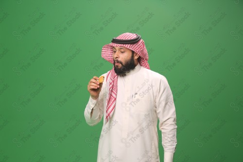 شاب سعودي على خلفية خضراء يرتدي الثوب و الشماغ، يقوم بعمل حركات بيده،يمسك الكعك و يأكله ، احتفالات العيد