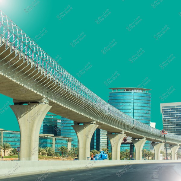 The image shows a modern overpass extending across the road, with tall glass buildings in the background. The sky is filled with a pure cyan color, and some palm trees are alongside the road. The architectural design of the buildings and the overpass reflects a contemporary style.