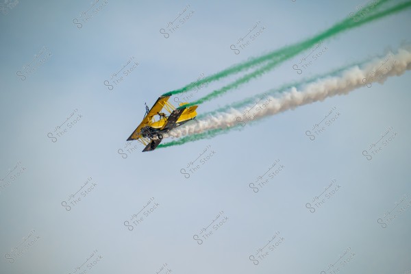 Image of a vintage biplane in yellow color, flying in the sky. The plane is emitting colored smoke in green and white behind it, indicating an aerial show. The background is a clear blue sky.