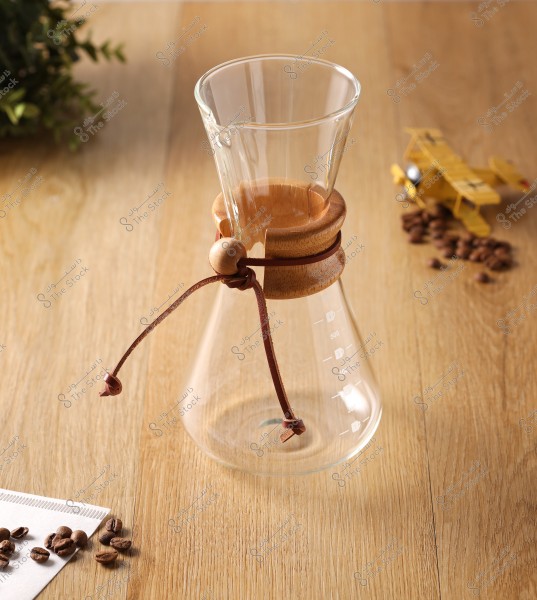 A glass coffee maker placed on a wooden table with scattered coffee beans and a small airplane model in the background.