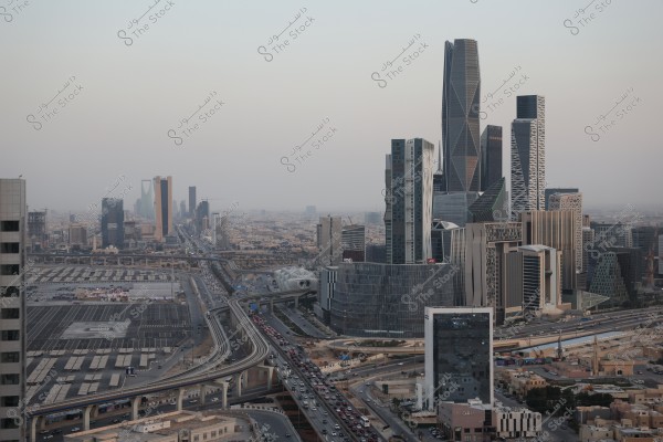 An aerial view of Riyadh, Saudi Arabia, showcasing a collection of modern skyscrapers with unique architectural designs. The image features several tall towers alongside highways bustling with cars. The weather appears slightly overcast, adding a layer of haze to the horizon.