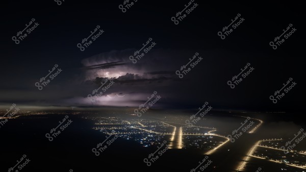 صورة ليلية لمدينة مضاءة بأنوار الشوارع, مع سماء مظلمة تحوي سحب كثيفة تضاء بالبرق في الأفق. تظهر الطرق مضاءة بشكل متقاطع, مع تجمعات صغيرة من الأضواء تشير إلى المناطق السكنية.