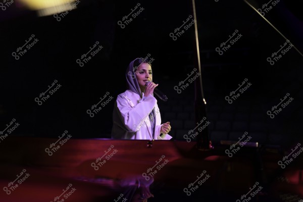 امرأة ترتدي حجابًا وتقف على المسرح تحمل ميكروفونًا, تبدو مضاءة بإضاءة مسرحية.