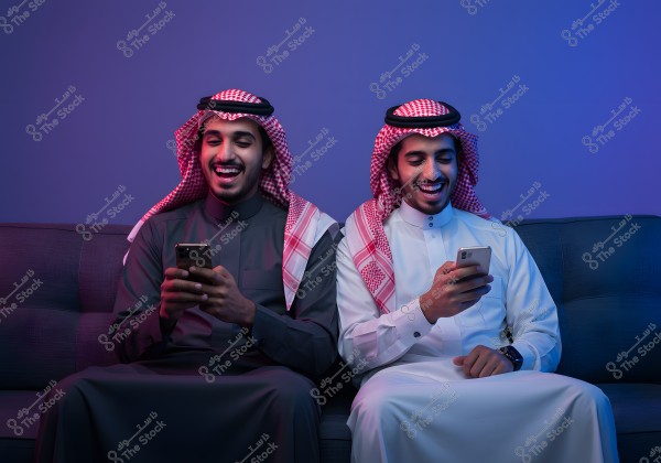 An image of two friends sitting on a sofa, wearing traditional Saudi attire. The person on the left is in a dark thobe with a red and white checkered headscarf, while the person on the right wears a white thobe with a similar headscarf. Both are smiling and looking at their smartphones, conveying a cheerful and friendly atmosphere.