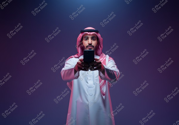 A portrait of a man wearing traditional attire, including a thobe and a ghutra, standing facing the camera while holding a smartphone in his hands. The background is lit with dark colors, adding a dramatic effect to the image. The clothing suggests a likely origin from the Gulf region, such as Saudi Arabia.