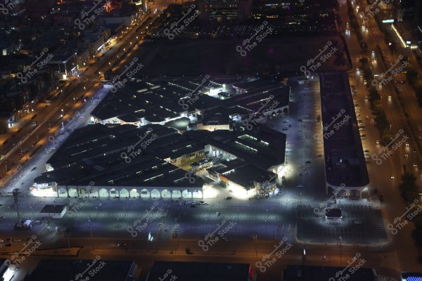 Aerial nighttime view of a city, showcasing beautifully lit streets and symmetrically designed buildings illuminated with bright night lights. The image features a large building with a sophisticated geometric design and a parking area surrounding it. The contrast between the bright lights and deep shadows in the surrounding area adds depth and aesthetic appeal to the image.